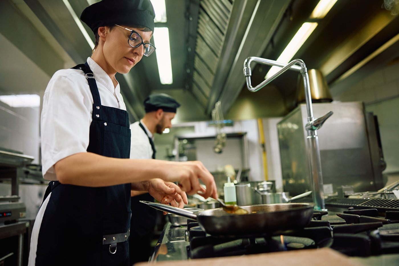 A professional chef in a white shirt and dark navy apron cooking in a commercial kitchen, stirring a pan on a stove while wearing glasses.