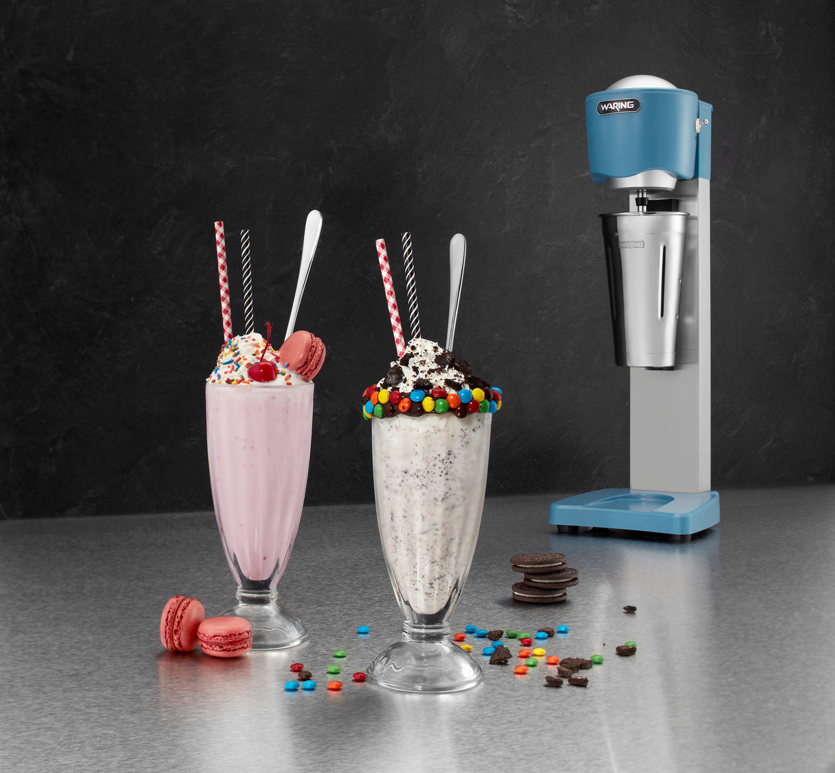 Two milkshakes—one pink with sprinkles and a macaron, the other cookies & cream with mini chocolate candies—sit on a counter with straws and spoons. Behind them is a blue Waring WDM20 Single-Spindle Drink Mixer (9x10-3/8x22, 2-speed manual).