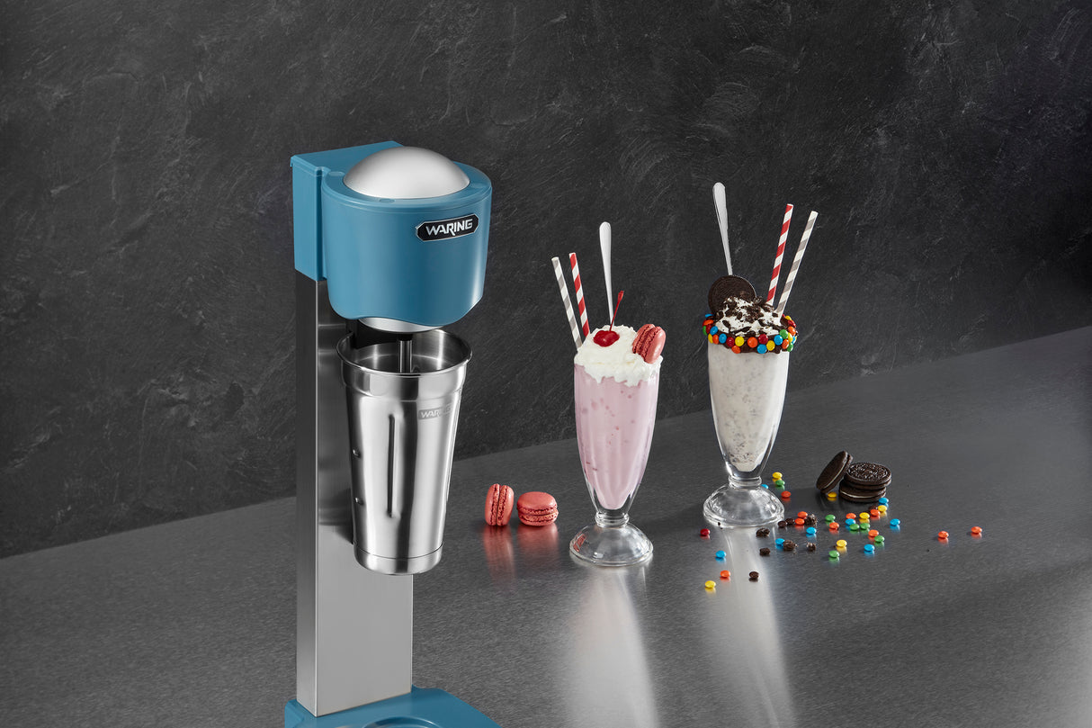 A Waring WDM20 Light-Duty Single-Spindle Drink Mixer stands on a countertop beside two tall milkshakes topped with whipped cream, cookies, macarons, and colorful candies. The background features a dark textured wall.